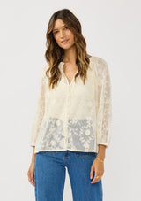 [Color: Natural] A front facing image of a brunette model wearing a sheer lace blouse with floral embroidery. Featuring a classic button up design, split v-neckline, and long sleeves. A breezy top with no lining for warm weather styling. 