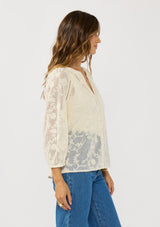 [Color: Natural] A side facing image of a brunette model wearing a sheer lace blouse with floral embroidery. Featuring a classic button up design, split v-neckline, and long sleeves. A breezy top with no lining for warm weather styling. 