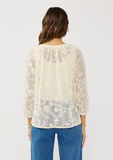[Color: Natural] A back facing image of a brunette model wearing a sheer lace blouse with floral embroidery. Featuring a classic button up design, split v-neckline, and long sleeves. A breezy top with no lining for warm weather styling. 