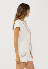 [Color: White/Dusty Rose] A side facing image of a brunette model wearing a white short sleeve top with pink embroidery, lace inserts, and a split v-neckline. Styled with matching bottoms. 
