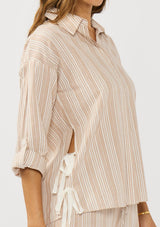[Color: Sand/White] A side facing image of a brunette model wearing a striped shirt with a collared neckline, button front, roll tab sleeves, and a playful side tie detail. 