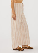 [Color: Sand/White] A side facing image of a brunette model wearing a beige striped bottom with wide legs and a an elastic drawstring waist.
