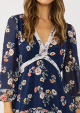 [Color: Navy/Coral] A detail image of a brunette model wearing a navy floral dress with an empire waist, v-neckline, long sleeves, and a tiered flowy skirt. A feminine flowy chiffon dress for the spring season. 