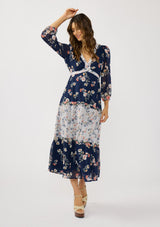 [Color: Navy/Coral] A front facing image of a brunette model wearing a navy floral dress with an empire waist, v-neckline, long sleeves, and a tiered flowy skirt. A feminine flowy chiffon dress for the spring season. 