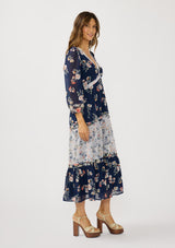 [Color: Navy/Coral] A side facing image of a brunette model wearing a navy floral dress with an empire waist, v-neckline, long sleeves, and a tiered flowy skirt. A feminine flowy chiffon dress for the spring season. 