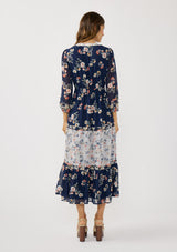 [Color: Navy/Coral] A back facing image of a brunette model wearing a navy floral dress with an empire waist, v-neckline, long sleeves, and a tiered flowy skirt. A feminine flowy chiffon dress for the spring season. 