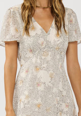 [Color: Natural/Dusty Peach] A detail image of a brunette model wearing a dotted floral chiffon midi dress with clipped lurex for a touch of sparkle. Featuring a flattering v-neckline, short flutter sleeves, and an elastic waist at the back. 