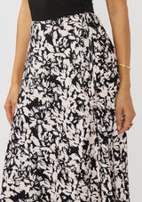 [Color: Vanilla/Black] A detail image of a brunette model wearing a black and white floral print skirt with a subtle pink tint, featuring long waist ties for an effortless wrap silhouette and a chic side tie. Lightweight and flowy, perfect for vacation or everyday wear.
