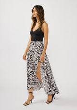 [Color: Vanilla/Black] A side facing image of a brunette model wearing a black and white floral print skirt with a subtle pink tint, featuring long waist ties for an effortless wrap silhouette and a chic side tie. Lightweight and flowy, perfect for vacation or everyday wear.