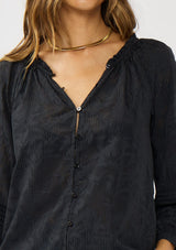 [Color: Black] A detail image of a brunette model wearing a black floral jacquard blouse crafted in a lightweight cotton blend. Featuring a chic button front, 3/4 length sleeve, and pintuck details. 