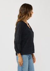 [Color: Black] A side facing image of a brunette model wearing a black floral jacquard blouse crafted in a lightweight cotton blend. Featuring a chic button front, 3/4 length sleeve, and pintuck details. 