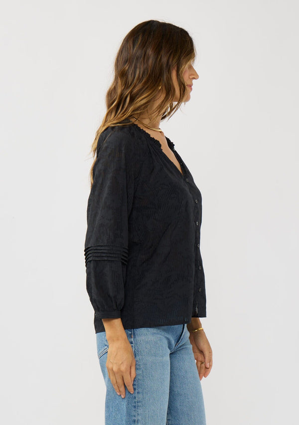 [Color: Black] A side facing image of a brunette model wearing a black floral jacquard blouse crafted in a lightweight cotton blend. Featuring a chic button front, 3/4 length sleeve, and pintuck details. 