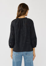 [Color: Black] A back facing image of a brunette model wearing a black floral jacquard blouse crafted in a lightweight cotton blend. Featuring a chic button front, 3/4 length sleeve, and pintuck details. 