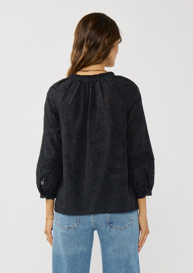 [Color: Black] A back facing image of a brunette model wearing a black floral jacquard blouse crafted in a lightweight cotton blend. Featuring a chic button front, 3/4 length sleeve, and pintuck details. 