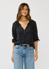 [Color: Black] A front facing image of a brunette model wearing a black floral jacquard blouse crafted in a lightweight cotton blend. Featuring a chic button front, 3/4 length sleeve, and pintuck details. 