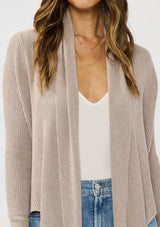 [Color: Heather Cement] A photo of a Lovestitch model wearing a chic beige waffle knit wrap front sweater with long sleeves, a v neckline, and a button closure at the back. The long ties can be styled in multiple ways.