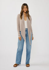 [Color: Heather Cement] A photo of a Lovestitch model wearing a chic beige waffle knit wrap front sweater with long sleeves, a v neckline, and a button closure at the back. The long ties can be styled in multiple ways.