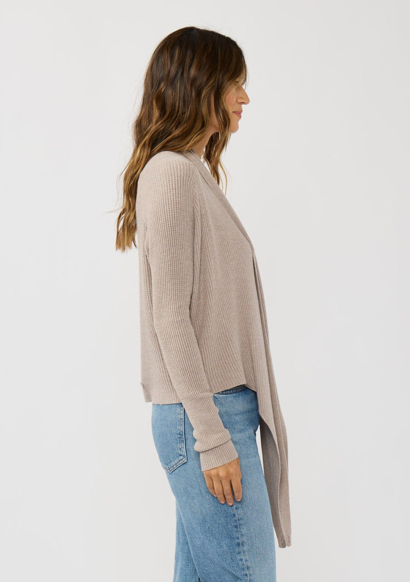 [Color: Heather Cement] A photo of a Lovestitch model wearing a chic beige waffle knit wrap front sweater with long sleeves, a v neckline, and a button closure at the back. The long ties can be styled in multiple ways.