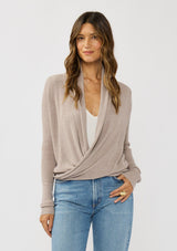 [Color: Heather Cement] A photo of a Lovestitch model wearing a chic beige waffle knit wrap front sweater with long sleeves, a v neckline, and a button closure at the back. The long ties can be styled in multiple ways.
