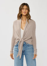 [Color: Heather Cement] A photo of a Lovestitch model wearing a chic beige waffle knit wrap front sweater with long sleeves, a v neckline, and a button closure at the back. The long ties can be styled in multiple ways.