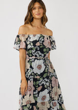 [Color: Black/Blush] A front facing image of a brunette model wearing a black and pink floral chiffon midi dress with an elastic off shoulder, braided waist belt, and tiered flowy skirt. A floral dress for brunch, spring styling, and special occasions. 