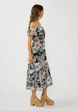 [Color: Black/Blush] A side facing image of a brunette model wearing a black and pink floral chiffon midi dress with an elastic off shoulder, braided waist belt, and tiered flowy skirt. A floral dress for brunch, spring styling, and special occasions. 