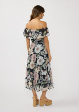 [Color: Black/Blush] A back facing image of a brunette model wearing a black and pink floral chiffon midi dress with an elastic off shoulder, braided waist belt, and tiered flowy skirt. A floral dress for brunch, spring styling, and special occasions. 