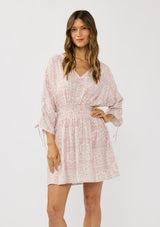 [Color: Vanilla/Rose] A front facing image of a brunette model wearing a pink floral print mini dress with long ruched sleeves with tie detail, a v-neckline, a smocked waist, and a partial button front. A casual bohemian mini dress for resort season.