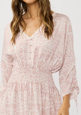 [Color: Vanilla/Rose] A detail image of a brunette model wearing a pink floral print mini dress with long ruched sleeves with tie detail, a v-neckline, a smocked waist, and a partial button front. A casual bohemian mini dress for resort season.