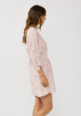 [Color: Vanilla/Rose] A side facing image of a brunette model wearing a pink floral print mini dress with long ruched sleeves with tie detail, a v-neckline, a smocked waist, and a partial button front. A casual bohemian mini dress for resort season.