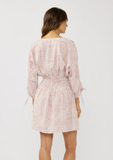 [Color: Vanilla/Rose] A back facing image of a brunette model wearing a pink floral print mini dress with long ruched sleeves with tie detail, a v-neckline, a smocked waist, and a partial button front. A casual bohemian mini dress for resort season.
