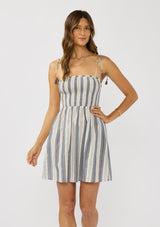 [Color: Blue/White] A front facing image of a brunette model wearing a cotton striped dress with a smocked bust, tie shoulder with tassels, and side pockets. An effortless coastal  mini dress for brunch or the beach. 