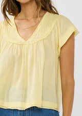 [Color: Yellow] A detail image of a brunette model wearing a flowy yellow bohemian top with short sleeves, a v-neckline, raw trim edges, and a shirred yoke. 