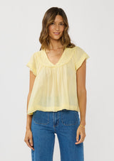 [Color: Yellow] A front facing image of a brunette model wearing a flowy yellow bohemian top with short sleeves, a v-neckline, raw trim edges, and a shirred yoke. 