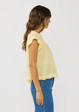 [Color: Yellow] A side facing image of a brunette model wearing a flowy yellow bohemian top with short sleeves, a v-neckline, raw trim edges, and a shirred yoke. 
