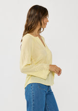 [Color: Yellow] A side facing image of a brunette model wearing a yellow blouse with tonal embroidery, long bell sleeves, and  split v-neckline. A lightweight and sheer top for the spring season. 