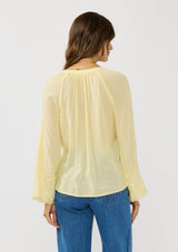 [Color: Yellow] A back facing image of a brunette model wearing a yellow blouse with tonal embroidery, long bell sleeves, and  split v-neckline. A lightweight and sheer top for the spring season. 