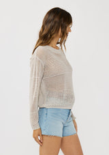 [Color: Stone] A side facing image of a brunette model wearing a grey knit top with silver metallic thread, a round neck, long sleeves, and open knit details. 