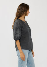 [Color: Grey] A side facing image of a brunette model wearing bohemian grey embroidered blouse with 3/4 length sleeves and a decorative button front. 