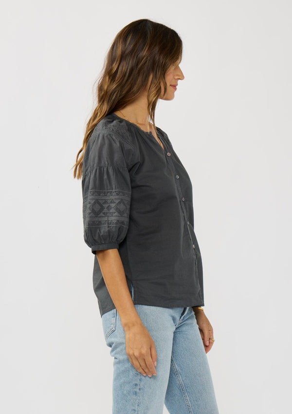[Color: Grey] A side facing image of a brunette model wearing bohemian grey embroidered blouse with 3/4 length sleeves and a decorative button front. 