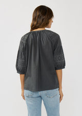 [Color: Grey] A back facing image of a brunette model wearing bohemian grey embroidered blouse with 3/4 length sleeves and a decorative button front. 