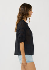 [Color: Black] A side facing image of a brunette model wearing an black sheer top with a split v-neckline and elbow length sleeves with pleats. A casual warm weather top for resort season. 