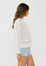 [Color: White] A side facing image of a brunette model wearing a plaid sheer blouse with lace and pintuck details. Featuring a split v-neckline, dainty tie neck detail, and long sleeves. 