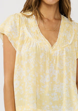 [Color: Yellow/Cream] A  detail image of a brunette model wearing a yellow floral print top with a v-neckline, shirred yoke, and short flutter sleeves. 