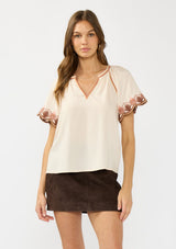 [Color: Sand] A front facing image of a brunette model wearing a light beige top with embroidered sleeves. Styled with a brown suede skirt. 