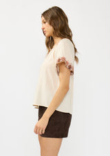 [Color: Sand] A side facing image of a brunette model wearing a light beige top with embroidered sleeves. Styled with a brown suede skirt. 