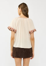 [Color: Sand] A back facing image of a brunette model wearing a light beige top with embroidered sleeves. Styled with a brown suede skirt. 