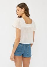 [Color: Natural/Cream] A back facing image of a brunette model wearing a white embroidered crop top with short flutter sleeves and a square neckline with tie detail.