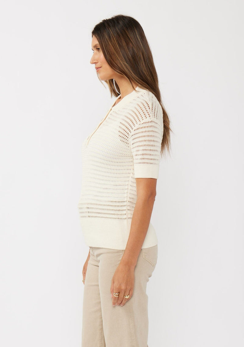 [Color: Vanilla] A side facing image of a brunette model wearing an off white cotton open knit top with short sleeves, ribbed trim, and a button front Henley placket.