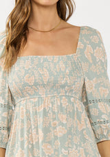 [Color: Dusty Sage/Natural] A detail image of a brunette model wearing a green floral maxi dress with a mocked bust, three quarter sleeves, and a square neckline. 